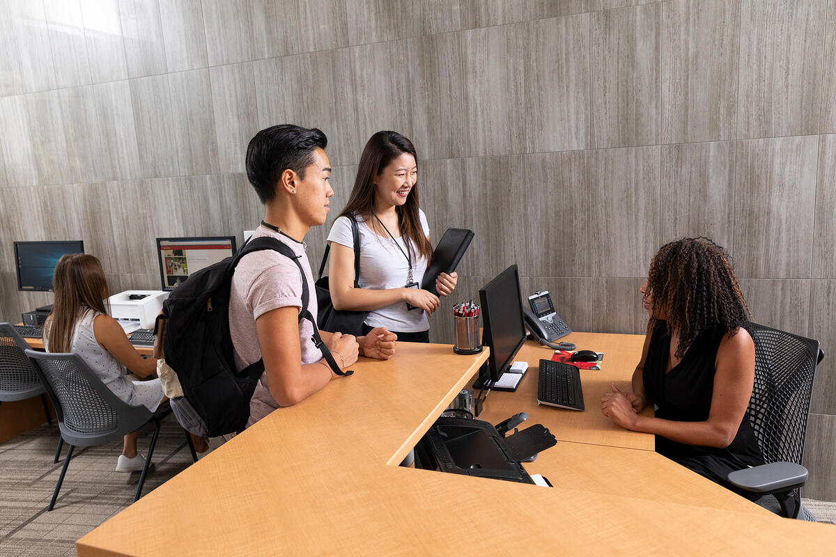 Two students talking with staff at a desk.
