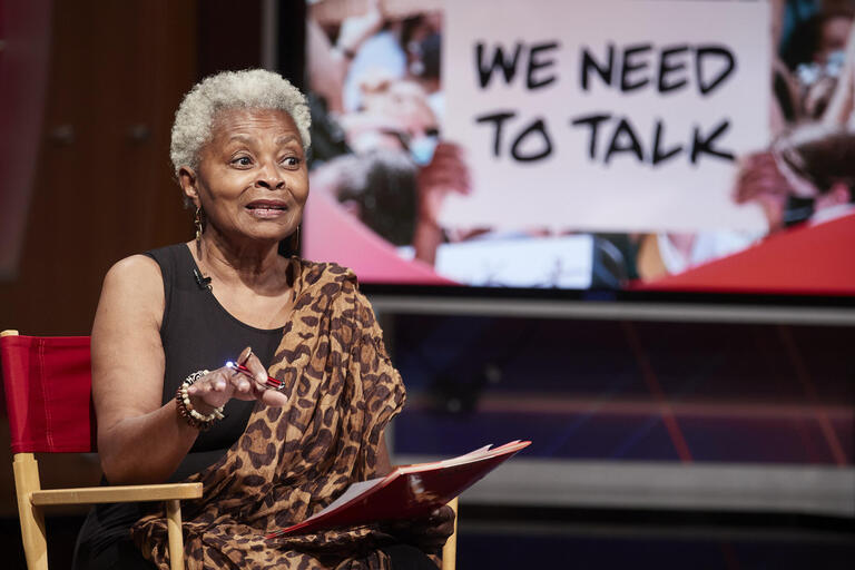 Claytee White seated with &quot;We Need to Talk&quot; on TV screen behind her