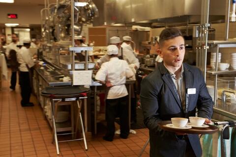 Young man carrying a tray of bowls out of a busy kitchen